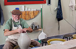 Potter wearing flat cap sitting at pottery wheel shaping clay vase