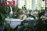 Nude young woman, sitting in bathtub, in bathroom full of plants, rear view