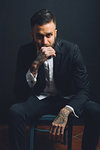 Portrait of young man, sitting on chair, wearing suit, tattoos on hands, pensive expression