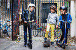 Boys riding push scooter and skateboard, bicycles in background