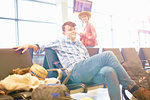 Young man sitting at airport, backpack beside him, young woman standing nearby, looking towards him
