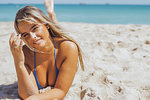 Pretty women in bikini and with long blond hair lying on sandy coast in tropical sunlight smiling alluringly at camera.