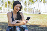 Attractive brunette in sportive clothing sitting in park using smartphone and smiling happily.