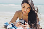 Content young woman with flying hair browsing smartphone and smiling while lying on beach and sunbathing.