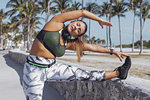 Attractive young woman in sportswear standing and stretching to legs while leaning on fence in the park.