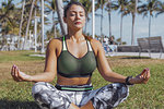 Athletic young woman in sportswear sitting with eyes closed and practicing yoga on green lawn in the park.