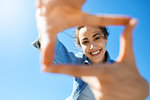 portrait of a young smiling attractive woman in jeans clothes at sunny day on the blue sky background. woman shows a frame from hands like photo. Photo Frame Hands Made By A Hipster young girl