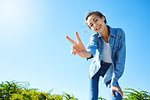 portrait of a young smiling attractive woman in jeans clothes at sunny day on the blue sky background. woman shows a Victory sign to the camera