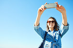 young smiling attractive woman in jeans clothes makes selfie on smartphone at sunny day on the blue sky background. woman posing in city scape.