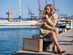 Attractive Young Blonde Woman in Trench and High Heel Shoes with Vintage Suitcase is Sitting on the Jacht Pier Looking in the Sea