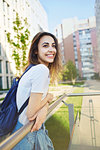 portrait of a young smiling attractive woman in white t-shirt with small city backpack at sunny day on city building background. woman poses in cityscape.