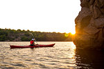 Young Woman Paddling the Red Kayak on the Beautiful River or Lake near High Rocks at Sunset