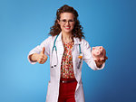 happy paediatrist woman in white medical robe with piggy bank showing thumbs up against blue background