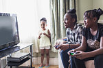 Toddler girl watching tween sisters playing video game in living room