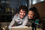 Happy couple using laptop and drinking white wine at home at night