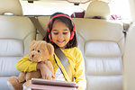 Smiling girl with teddy bear using digital tablet with headphones in back seat of car