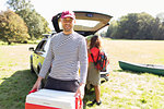 Portrait smiling man carrying camping cooler, unloading car in sunny field