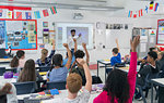 Male teacher leading lesson at projection screen in classroom with students raising hands