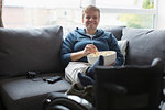 Smiling young woman watching TV and eating popcorn on sofa with feet up on wheelchair
