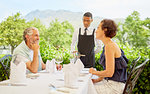 Wine steward pouring wine for mature couple dining at patio table