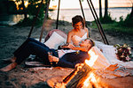 Bride and groom reclining by campfire on lakeside at dusk