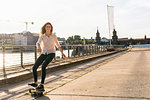 Young woman skateboarding on bridge, river and buildings in background, Berlin, Germany