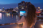 Young woman taking photograph with digital tablet on bridge, river and city in background, Berlin, Germany
