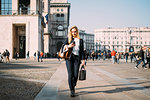 Young female tourist with shopping bags strolling and looking at smartphone in city square, Milan, Italy