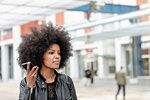 Young woman with afro hair at city train station, listening to smartphone