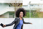 Happy young woman with afro hair in city, laughing and dancing to smartphone music