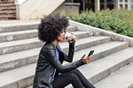 Young woman with afro hair sitting on city stairway, drinking takeaway coffee and looking at smartphone