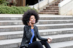 Happy young woman with afro hair sitting on city stairway, listening to smartphone