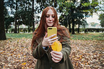 Young woman with long red hair in park taking smartphone photo of her hand holding autumn leaf
