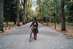 Young woman with long red hair pushing bicycle in tree lined  autumn park, Florence, Tuscany, Italy