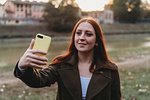 Young woman with long red hair taking smartphone selfie on riverside at dusk, Florence, Tuscany, Italy