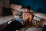 Mid adult man with tattooed chest on sofa looking at laptop