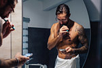 Mid adult man with tattoos brushing teeth and looking at smartphone at bathroom mirror, mirror image