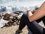 Young female surfer sitting on beach rock amongst  ocean waves, cropped, Cape Town, Western Cape, South Africa