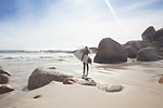 Young female surfer carrying surfboard looking out at ocean waves from beach, full length, Cape Town, Western Cape, South Africa