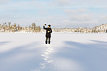 Woman taking photograph with smart phone on snow covered Stora Skiren lake in Lotorp, Sweden