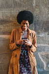 Young woman with afro hair using smartphone, leaning against stone wall