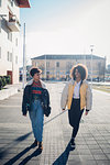 Two cool young female friends strolling on urban sidewalk, full length