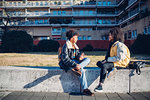 Two cool young female friends sitting on urban wall chatting
