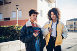 Two cool young female friends with smartphones on urban sidewalk, portrait