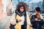 Two cool young women strolling and looking at smartphone on urban sidewalk