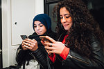 Young woman in hijab and friend sitting on subway train looking at smartphones