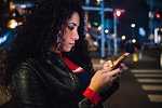 Mid adult woman with long curly hair looking at smartphone on city street at night