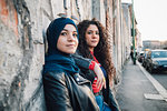 Young woman in hijab and best friend leaning against wall on city sidewalk