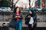 Young woman in hijab and her friend using smartphones and laughing by city canal