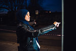 Young woman in hijab using illuminated touchscreen at city bus stop at night